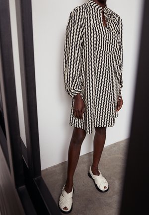 Personne portant une robe mi-longue à rayures ondulées noires et blanches et des sandales tissées blanches, debout sur un sol gris contre un mur blanc.