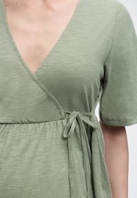 Γυναίκα που φοράει ανοιχτοπράσινο κοντομάνικο τοπ με ντραπέ V-λαιμόκοψη και δεσίματα στο πλάι, κομμένο στο στήθος και στη μέση.