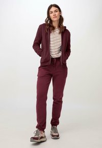 Burgunderfarbene Kapuzenjacke mit Fronttaschen und Reißverschluss, kombiniert mit passenden, tapered Jogginghosen. Getragen über einem gestreiften, kurzärmeligen Oberteil. Neutrale Turnschuhe.