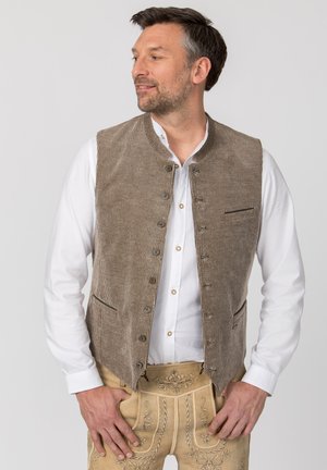 Mann trägt weißes Hemd, braun gestreifte Weste und beige bestickte traditionelle Hose, schaut nach links, Hände auf den Hüften.