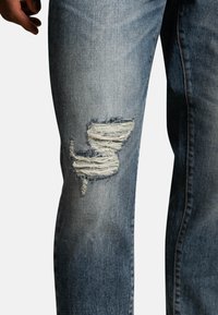Light blue distressed jeans featuring a large, frayed hole on the left knee, with a textured denim finish and subtle fading.