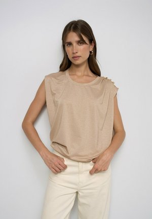 Mujer con pelo largo y castaño que lleva una camiseta beige sin mangas con botones en un hombro y pantalones de color claro, de pie frente a un fondo sencillo.