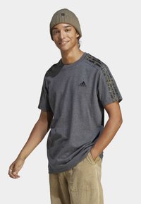 Siv majica s črnim logotipom Adidas, poudarjena z rameni s kamuflažnim vzorcem. Oblečena z bež kordnimi hlačami, model se smeška med poziranjem.