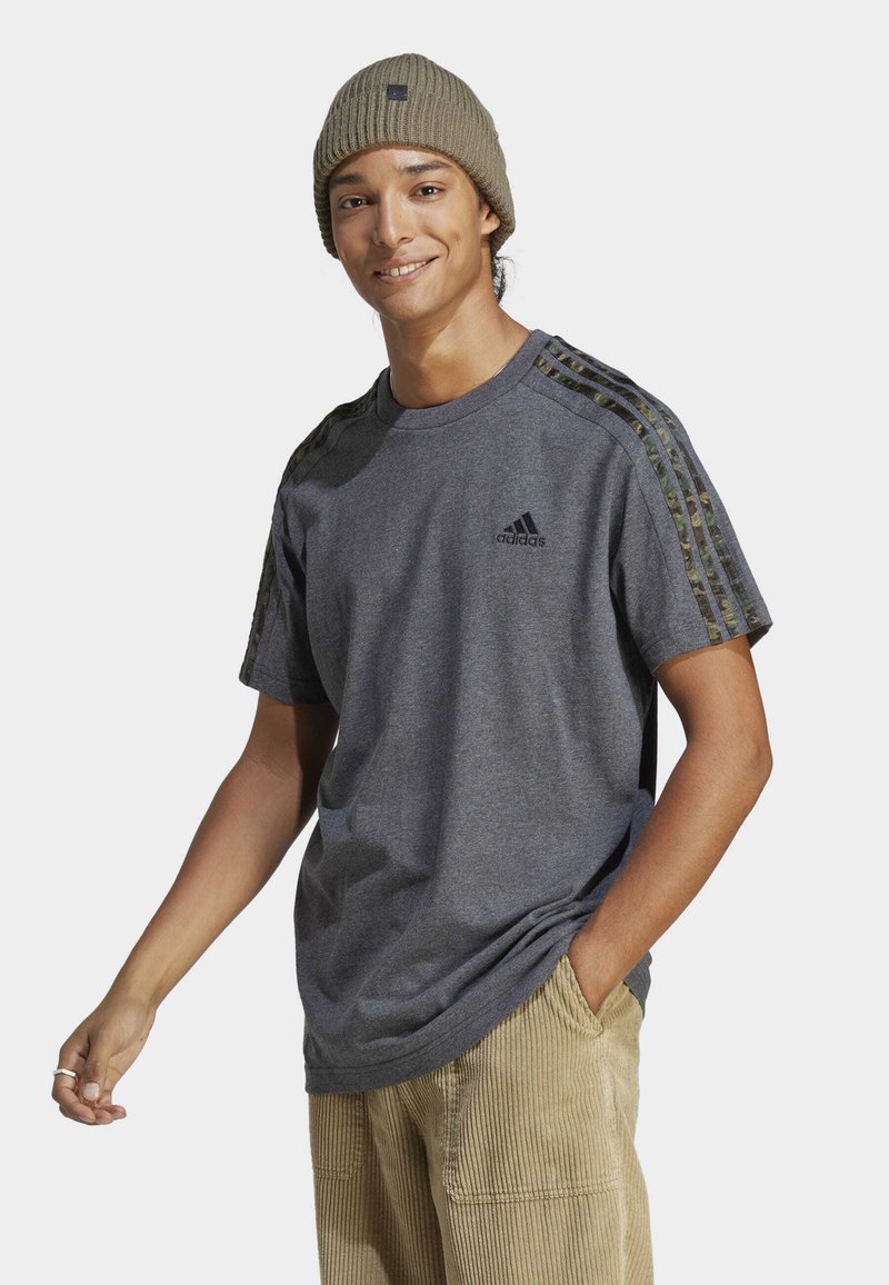 Siv majica s črnim logotipom Adidas, poudarjena z rameni s kamuflažnim vzorcem. Oblečena z bež kordnimi hlačami, model se smeška med poziranjem.