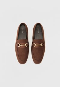 Brune suedé loafers med en strukturert design, som har en gullmetallaccent og elastisk bånd. Myk tekstur med rund tåform.