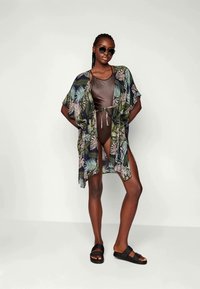 Kimono ligero y abierto con un estampado de hojas tropicales en verde y crema, combinado con un traje de baño marrón y sandalias negras.