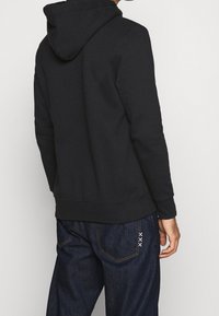 Sudadera con capucha negra de tela suave y corte holgado, con puños y dobladillo ribeteados. Combinada con vaqueros oscuros que tienen costuras decorativas.