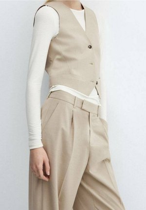 Vrouw draagt beige vest en beige broek met hoge taille over een wit top met lange mouwen, staand tegen een effen lichte achtergrond.