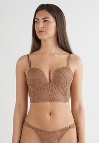 Koronkowy bralet w bogatym odcieniu brązu z floralnym wzorem, z dekoltem w serek i regulowanymi ramiączkami. Pasujące koronkowe stringi wyeksponowane poniżej.