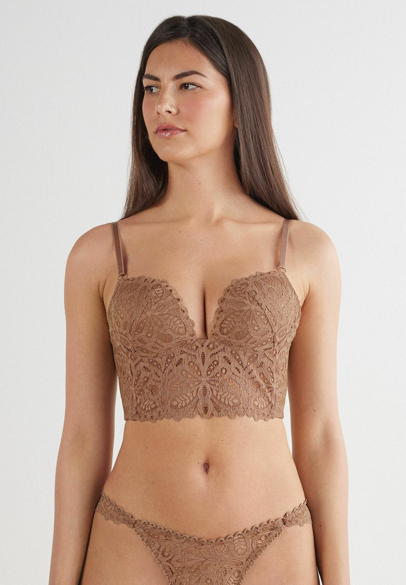 Koronkowy bralet w bogatym odcieniu brązu z floralnym wzorem, z dekoltem w serek i regulowanymi ramiączkami. Pasujące koronkowe stringi wyeksponowane poniżej.