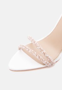 Άσπρο μυτερό νυφικό παπούτσι με ανοιχτά δάχτυλα και ψηλό τακούνι, με δύο λουράκια διακοσμημένα με μαργαριτάρια και χάντρες στο μπροστινό μέρος.