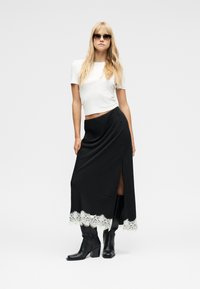 Ξανθιά γυναίκα με γυαλιά ηλίου, λευκό κοντό μπλουζάκι, μαύρη μακριά φούστα με λευκή δαντέλα στο τελείωμα και μαύρα μποτάκια με τακούνι, ποζάροντας σε λευκό φόντο.