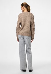 Beżowy sweter na drutach z fakturowanym wzorem, opadającymi ramionami i luźnym krojem, połączony z jasnoszarymi dżinsami o szerokich nogawkach.