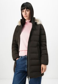 Kvinde med glat, mørkt hår iført en sort polstret frakke med hætte med pelskant, lyserød halskrave sweater og blå jeans, stående foran en hvid baggrund.