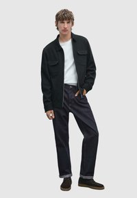 Veste zippée noire avec deux poches frontales sur une chemise blanche, associée à un jean en denim foncé et des chaussures marron. Fond gris clair.