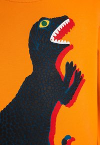 Camisa naranja con un gráfico de dinosaurio en azul y negro, con un patrón de escamas texturizado y acentos rojos alrededor de la boca y las garras.