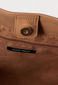 Interiér hnedej textilnej tašky s kovovým zacvakávacím zapínaním a vreckom na zips. Štítok nesie nápis „ANNA FIELD“. Mäkká, hladká textúra.