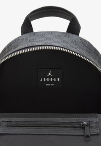 Sort rygsæk med en struktureret top, der har et mønster, sølvlynlås og et sort logopatch med teksten "JORDAN".