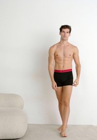 Boxers noirs avec une ceinture rose arborant le logo "BJORN BORG" ; modèle debout pieds nus, avec une peau lisse contre un fond neutre.