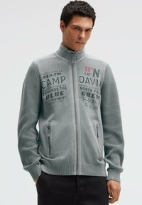 Grauer Pullover mit Reißverschluss und strukturiertem Strickmuster, versehen mit aufgedruckten Textdetails, hohem Kragen und Seitentaschen. Schlanke Passform.