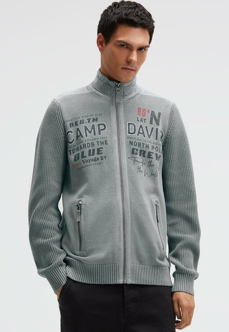 Grauer Pullover mit Reißverschluss und strukturiertem Strickmuster, versehen mit aufgedruckten Textdetails, hohem Kragen und Seitentaschen. Schlanke Passform.