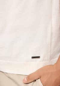 Maglione lavorato a maglia color beige chiaro con orlo a coste, dotato di un'etichetta del marchio in metallo nero in basso a sinistra. Tessuto morbido e vestibilità casual.