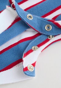 Tkanina w paski niebieskie, białe i czerwone z częściowo zapiętymi srebrnymi zatrzaskami na dzianinowym ubraniu, pokazana na jasnej powierzchni.