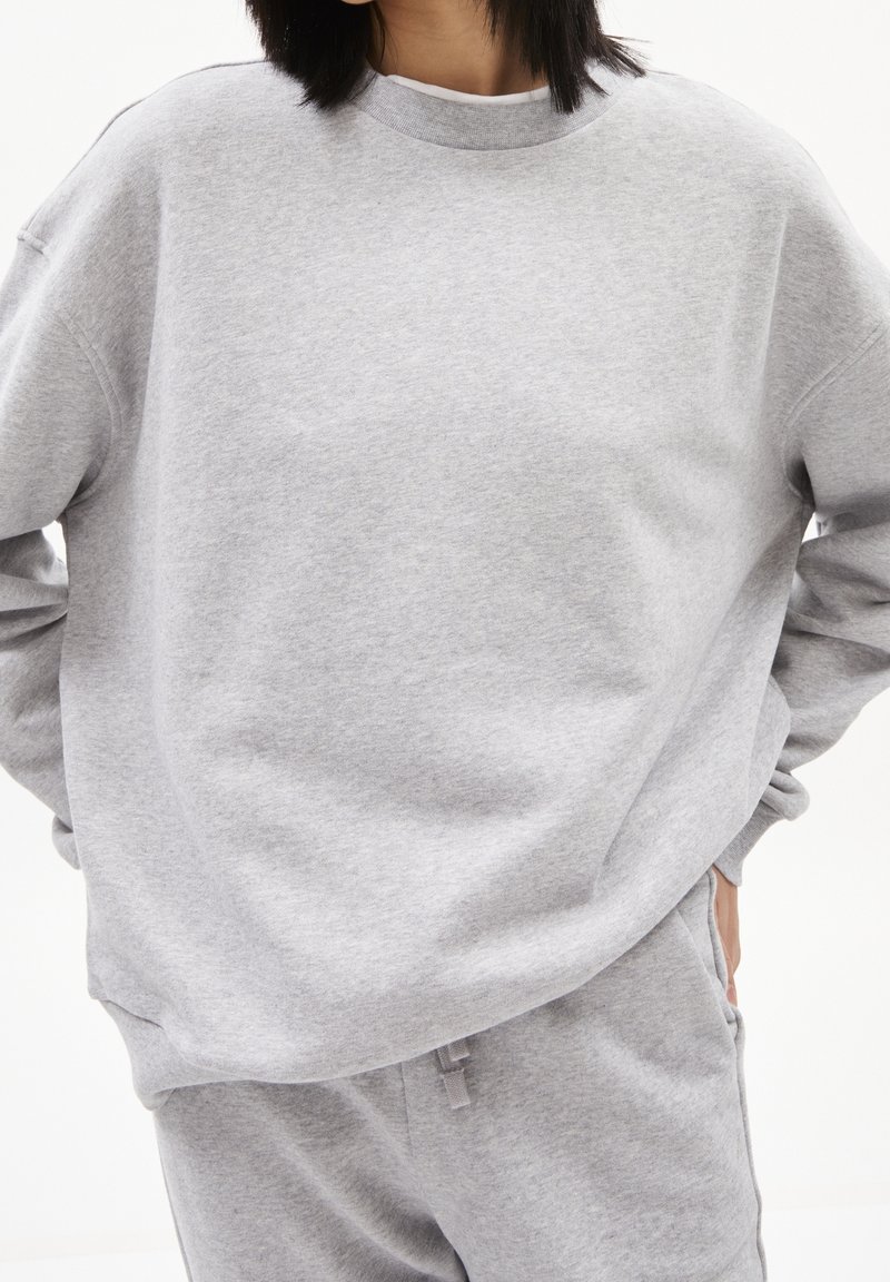 Personne portant un sweat-shirt gris clair ample et un pantalon de jogging assorti, les mains dans les poches sur fond blanc.