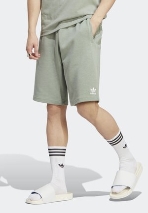 Osoba nosząca jasnospodenki Adidas w kolorze jasnozielonym, białe skarpetki Adidas z czarnymi paskami oraz białe klapki na biegu, idąca z jedną ręką w kieszeni.