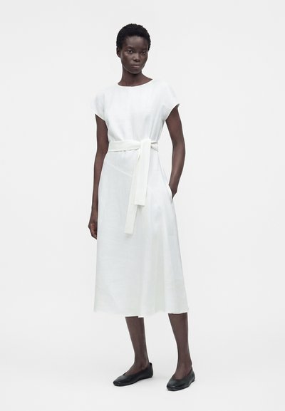 Γυναίκα που φοράει λευκό midi φόρεμα με κοντά μανίκια και δεμένη ζώνη στη μέση, συνδυασμένο με μαύρα επίπεδα παπούτσια, στέκεται μπροστά σε λευκό φόντο.