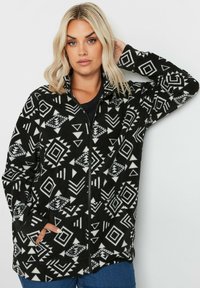 Giacca in fleeces con una base nera e motivi geometrici bianchi. Presenta una chiusura con zip e tasche laterali. Il tessuto appare morbido e texturizzato.