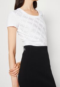 Mujer vistiendo una blusa blanca de manga corta con patrones de mariposas plateadas y una falda negra de cintura alta con un pequeño logo "CK" cerca de la cintura.