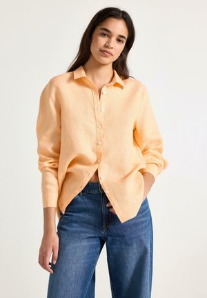 Kvinde med langt mørkt hår iført en lys orange skjorte med knapper og blå jeans, stående med den ene hånd i lommen mod en ensfarvet baggrund.