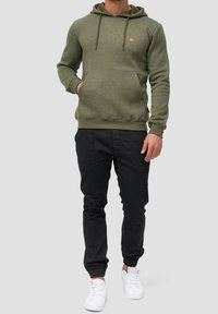 Sudadera verde oliva con un bolsillo tipo canguro y cordones. Pantalones negros con puños elásticos. Zapatillas blancas. Diseño sencillo y enfocado en la comodidad.
