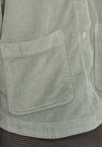 Tasca di giacca di velluto a coste verde chiaro con cuciture visibili e un bottone argentato sopra, abbinata a pantaloni a quadri scuri sotto.