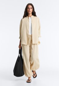 Femme portant un pantalon beige ample texturé et une chemise boutonnée assortie, un haut blanc, des sandales noires, tenant un grand sac fourre-tout noir, marchant vers l’avant.