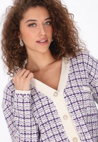 Lila és fehér kötött kardigán V-nyakkal, textúrázott mintázattal és díszes gombokkal. Jellemzője a bordázott szegély az ujjakon és a galléron.