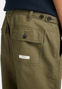 Olijfgroene cargobroek met twee achterzakken, donkere knopen en een label met de tekst "ELEMENT CO. SINCE 1992." Stevige stof met een soepele textuur.