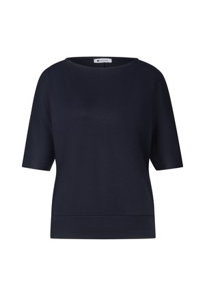 Femme aux cheveux châtain mi-longs souriante, portant un haut ample bleu marine foncé à manches courtes sur un fond uni.