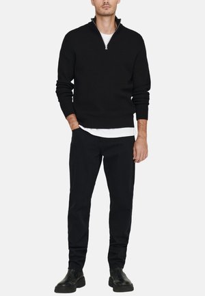 Zwarte gebreide trui met een ritskraag, gecombineerd met zwarte broek en zwarte schoenen. De outfit heeft een gelaagd wit shirt eronder.