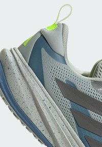 Vaaleanharmaa urheilujalkine sinisillä yksityiskohdilla, rakenteellinen verkkopinta, näkyvät harmaat raidat ja neonvihreä vetotuli. Varustettuna rosoisella pohjalla.