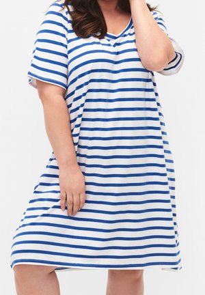 Vrouw met een witte jurk tot op de knie met horizontale blauwe strepen, korte mouwen en een V-hals, staand tegen een effen achtergrond.