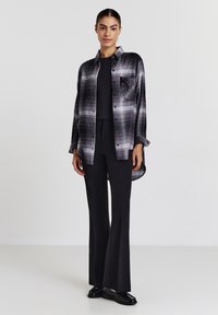 Schwarz-grau karierte Hemd mit Tasche und dekorativen Nieten, getragen über einem schwarzen Oberteil und kombiniert mit enganliegenden schwarzen Hosen und Schuhen.