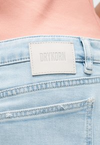 Lyseblå denimjeans med en hvit sydd lapp med teksten «DRYKORN» på linningen, brukt sammen med en rosa ribbestrikket topp.
