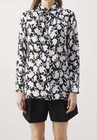 Camisa de flores en blanco y negro de manga larga con botones en la parte delantera y detalle de cuello fruncido, de tela transparente, combinada con pantalones cortos negros.