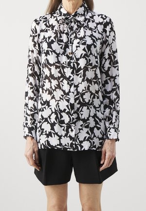 Schwarzes und weißes florales Langarmhemd mit durchgehender Knopfleiste und gerafftem Kragen, aus durchsichtigem Stoff, getragen mit schwarzen Shorts.