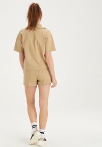 Tan polo tričko s krátkým rukávem a embosovaným textem, spárované s odpovídajícími šortkami. Hladká tkanina, neformální design, nošeno s atletickými botami a pruhovanými ponožkami.
