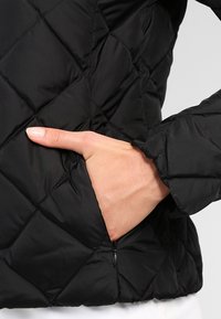 Veste matelassée noire avec une poche zippée sur le côté, présentant une texture lisse, un design structuré et un motif en losanges dans le tissu.