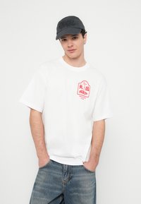 Bílé bavlněné tričko s červeným potiskem na levé straně, v kombinaci s modrými džínami a tmavou baseballovou čepicí. Neformální střih.