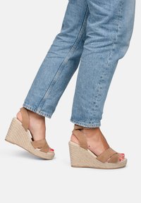 Sandali a zeppa in suede marrone con suola in juta intrecciata, dotati di due fasce incrociate e una cinghia alla caviglia, abbinati a jeans in denim azzurro.